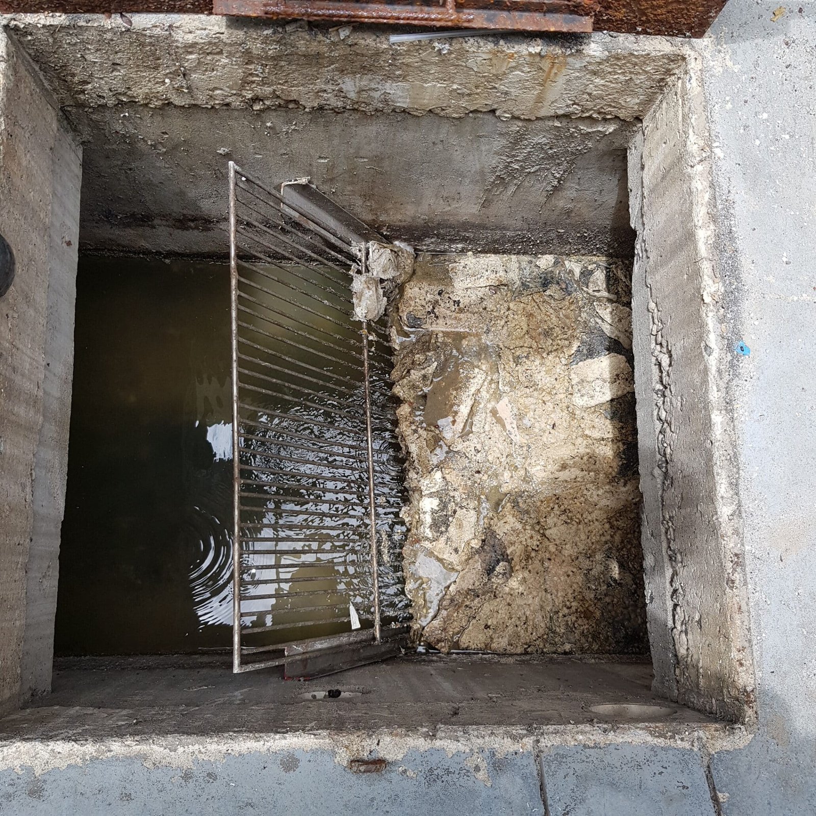 Cámara de acceso a pozo de alcantarillado con rejilla metálica y agua estancada en túnel subterráneo