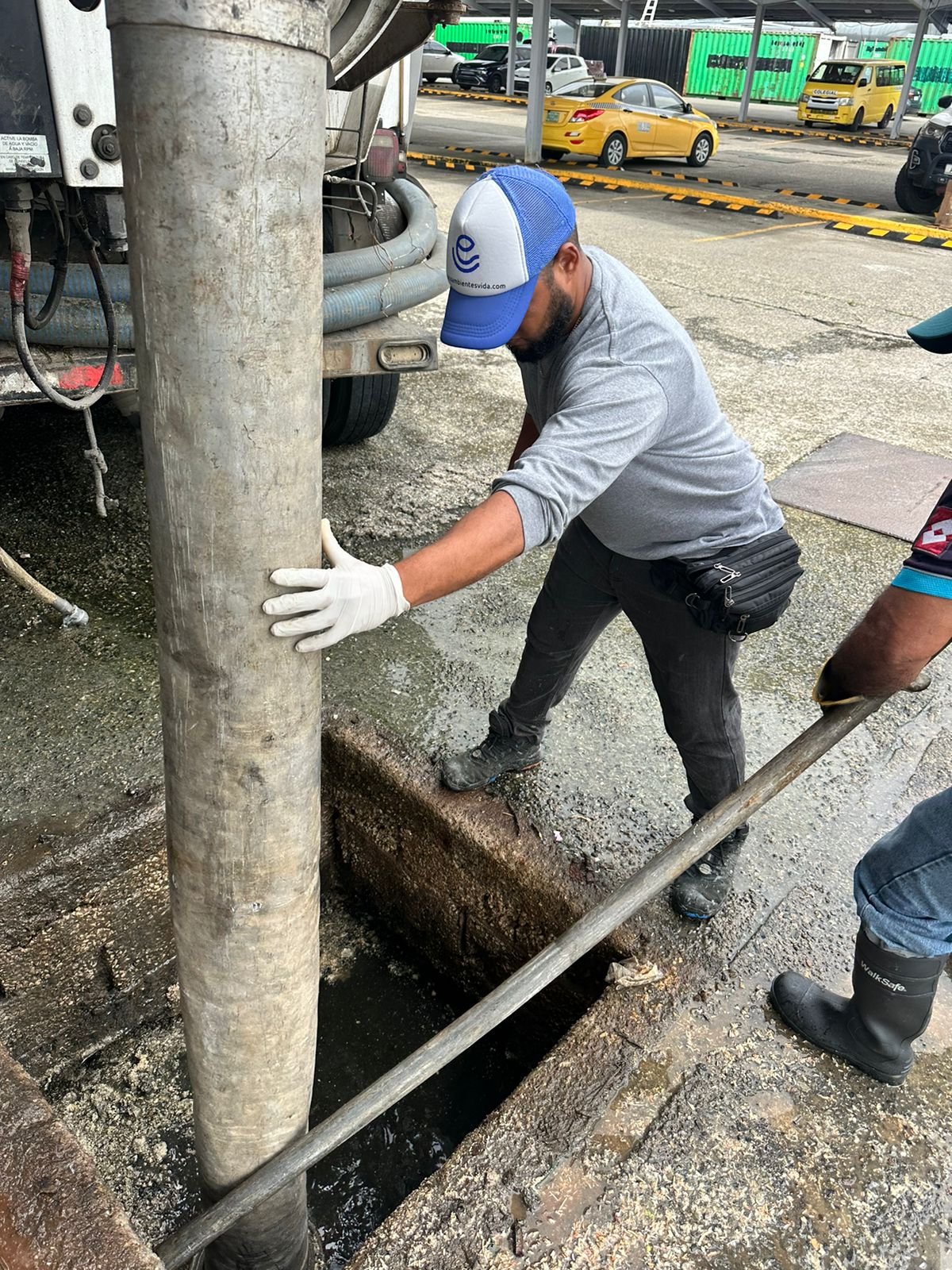 Operario en equipamiento de seguridad realizando mantenimiento de sistema de alcantarillado en calle urbana con camiones cisterna de fondo