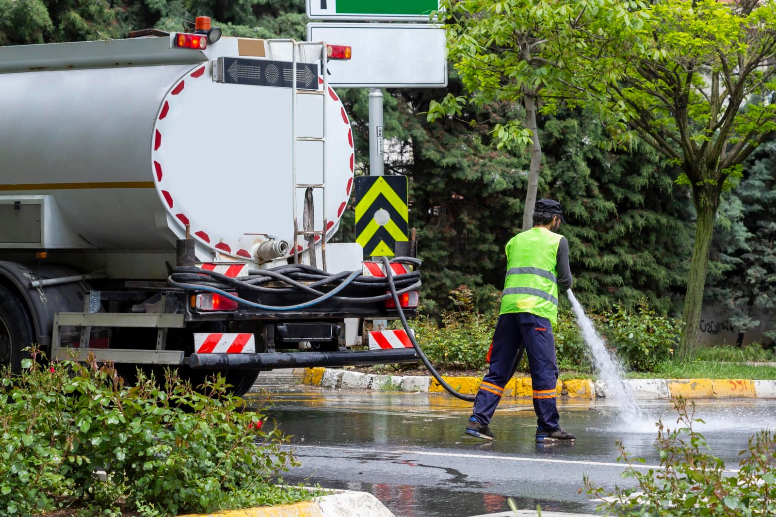 Camión cisterna de limpieza húmeda limpiando calle urbana con trabajador en equipo de seguridad amarillo y azul
